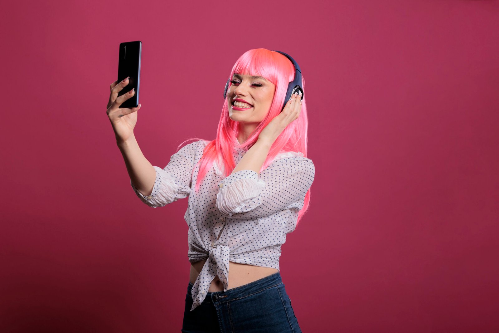 Female model with pink hair taking pictures or recording video on smartphone, listening to music on headphones. Using mobile phone to take photos and enjoy songs on headset for entertainment.