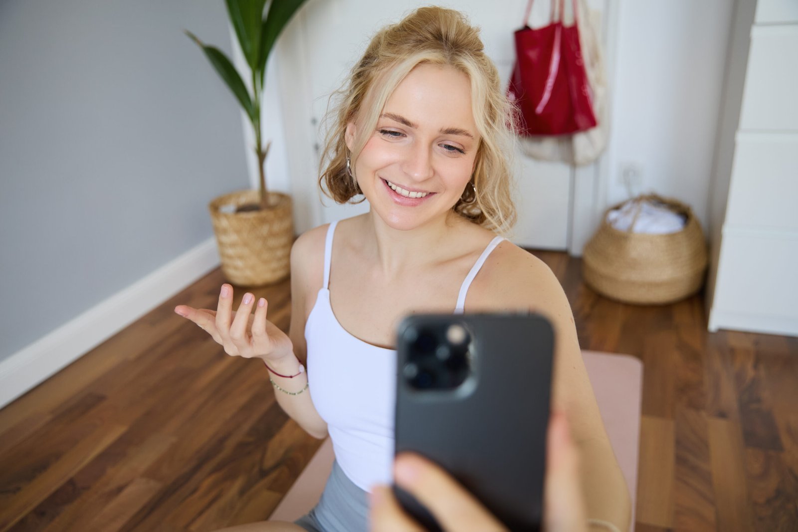 Portrait of beautiful young fitness trainer, showing home workout, taking selfie on mobile phone while sitting on yoga rubber mat in bedroom, wearing activewear.